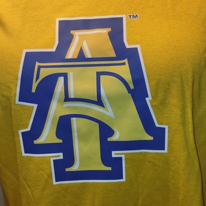 May include: A yellow t-shirt featuring a large, stylized "AT" logo in blue and yellow with a white outline. The shirt is made of a soft material and is displayed on a mannequin.