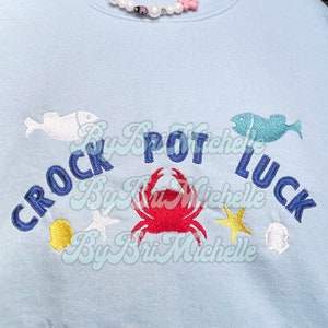 Może przedstawiać: Jasno błękitna bluza z haftowanym tekstem "Crock Pot Luck" w łuku, przedstawiającym czerwonego kraba, dwie białe ryby, żółtą muszlę, niebieską rybę i dwie złote rozgwiazdy.