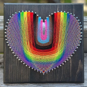 May include: A rainbow-colored string art heart on a dark wood background. The heart is made of colorful threads wrapped around nails.