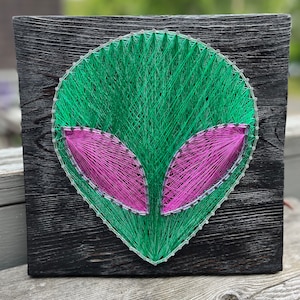 May include: Green and pink string art of an alien head on a dark wood background.