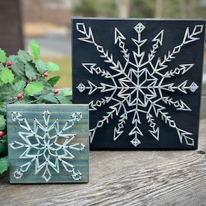 May include: Two wooden signs with string art snowflakes. One sign is black with a white snowflake, the other is blue with a white snowflake.