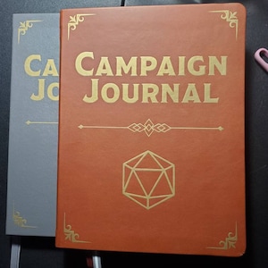 May include: Two brown leather-bound journals with gold accents. The journal on the right has the text "CAMPAIGN JOURNAL" in gold lettering and a gold-colored 20-sided die graphic.