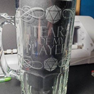 May include: A clear glass beer mug with a white etched design. The design includes a 20-sided die, a stylized knot, and the text "Tears of my Player".