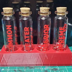 May include: Four clear glass bottles with red labels and red dice inside. The labels read "Common", "Greater", "Superior", and "Supreme". The bottles are sitting in a red plastic holder.