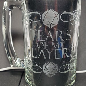 May include: A clear glass beer mug with a handle. The mug is etched with a 20-sided die, a decorative swirl, and the text "Tears of my players".