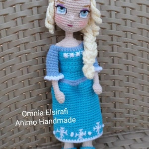 May include: Crocheted doll with blonde hair, wearing a blue dress with white snowflake accents and blue shoes. The doll is sitting on a wicker chair. The text "Omnia Elsirafi Animo Handmade" is visible below the doll.