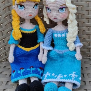May include: Two crocheted dolls dressed as the Disney princesses Elsa and Anna. Elsa is wearing a blue dress with snowflake embroidery and a white crocheted braid. Anna is wearing a blue dress with flower embroidery and a yellow crocheted braid. The dolls are sitting on a woven chair.