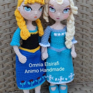 May include: Two crocheted dolls, one with blonde hair and a blue dress, the other with yellow hair and a blue and black dress. The dolls are standing on a woven surface. The text "Omnia Elsirafi Animo Handmade" is visible below the dolls.