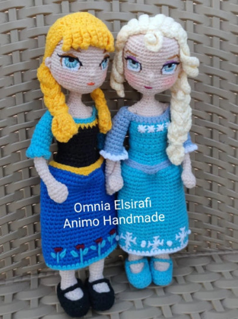 May include: Two crocheted dolls, one with blonde hair and a blue dress, the other with yellow hair and a blue and black dress. The dolls are standing on a woven surface. The text "Omnia Elsirafi Animo Handmade" is visible below the dolls.