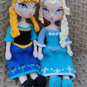 May include: Two crocheted dolls, one with blonde hair and a blue dress with flower accents, the other with white hair and a blue dress with snowflake accents. The dolls are sitting on a wicker surface. The text "Omnia Elsirafi Animo Handmade" is visible at the bottom of the image.