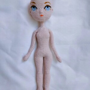 May include: A crocheted doll with a pink body, blue eyes, and orange lips. The doll is standing on a white surface.