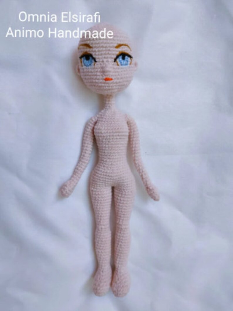 May include: A crocheted doll with a pink body, blue eyes, and orange lips. The doll is standing on a white surface.
