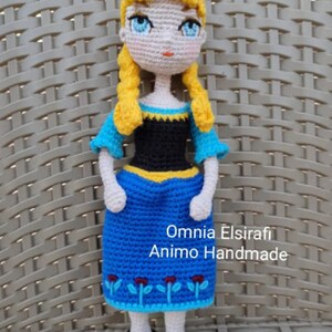 May include: A crocheted doll with blonde hair, blue eyes, and a blue and black dress. The doll is wearing black shoes and has a yellow and black trim on the dress. The doll is standing on a woven surface. The text "Omnia Elsirafi Animo Handmade" is visible below the doll.