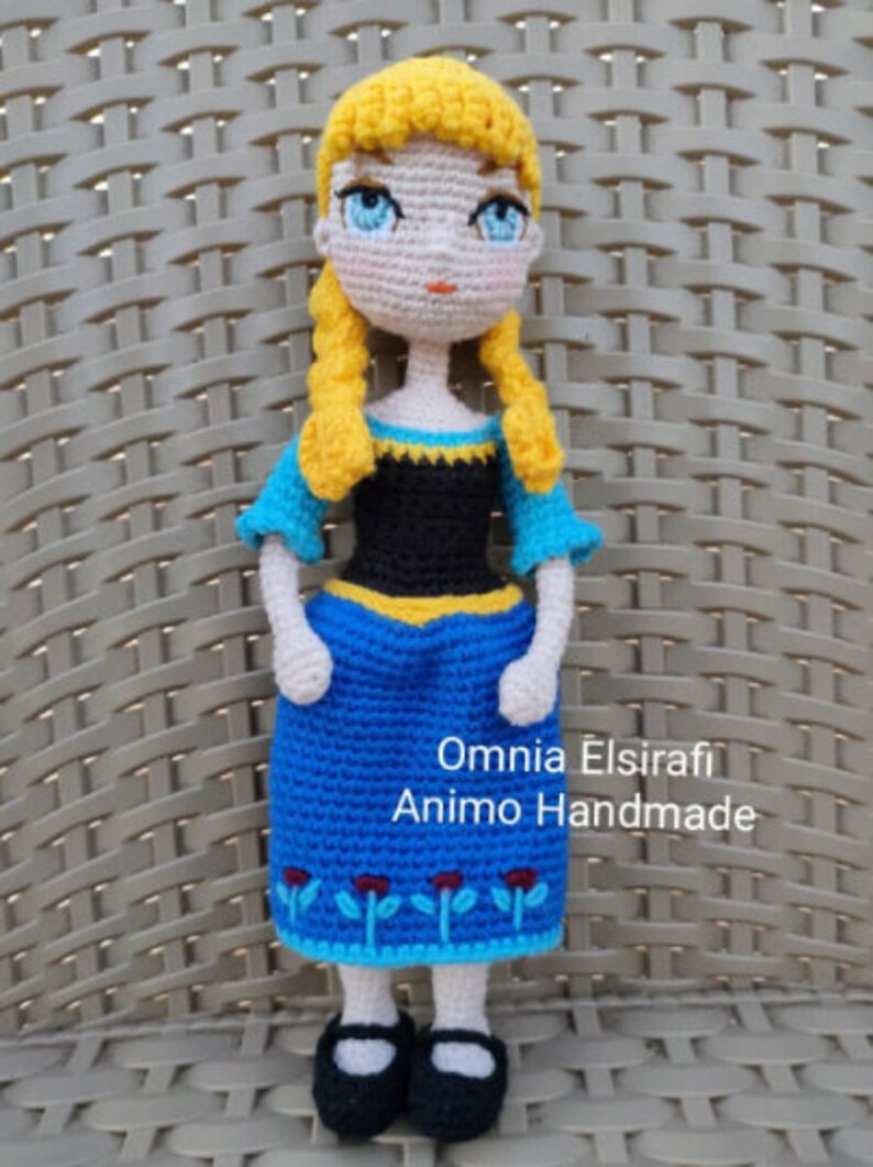 May include: A crocheted doll with blonde hair, blue eyes, and a blue and black dress. The doll is wearing black shoes and has a yellow and black trim on the dress. The doll is standing on a woven surface. The text "Omnia Elsirafi Animo Handmade" is visible below the doll.