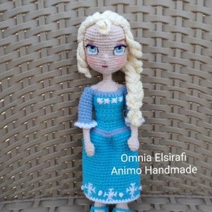 May include: A crocheted doll with blonde hair, wearing a blue dress with white snowflakes, and blue shoes. The doll is standing on a woven surface. The text "Omnia Elsirafi Animo Handmade" is visible below the doll.