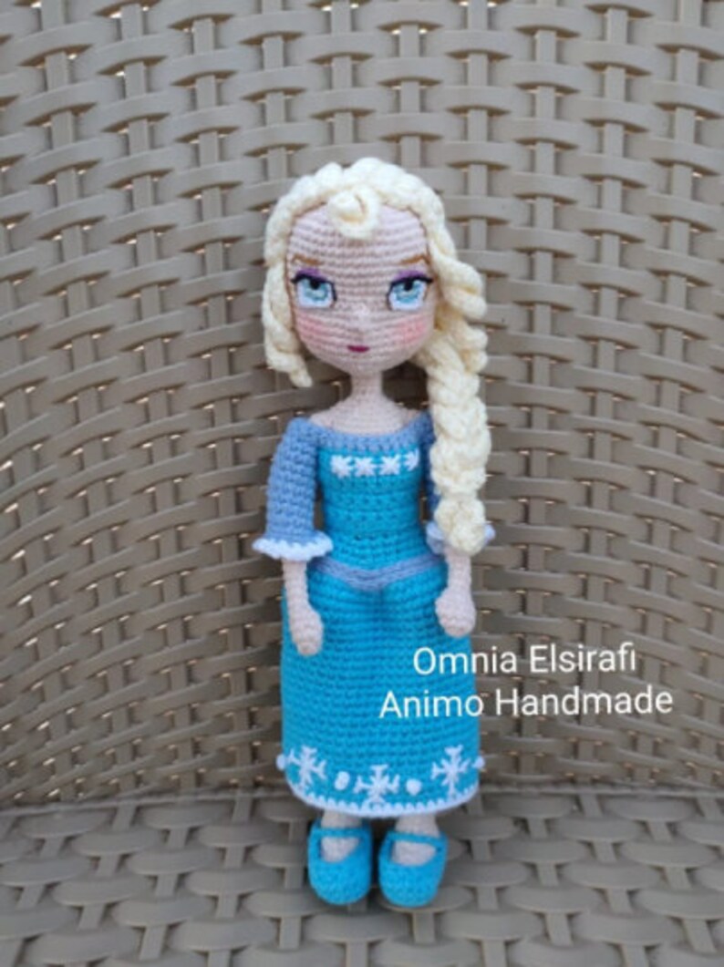 May include: A crocheted doll with blonde hair, wearing a blue dress with white snowflakes, and blue shoes. The doll is standing on a woven surface. The text "Omnia Elsirafi Animo Handmade" is visible below the doll.
