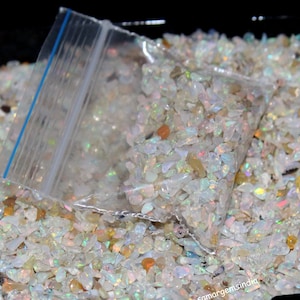May include: A transparent plastic bag containing small, iridescent opal chips, set on a bed of similar chips. The opals show flashes of colour, including white, yellow, and orange. The bag has a blue zip.