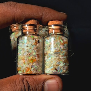 May include: Small glass bottles filled with colorful opal chips. Each bottle has a cork stopper. The opals display a variety of colors, including white, orange, and green. The bottles are held in a hand against a dark background.