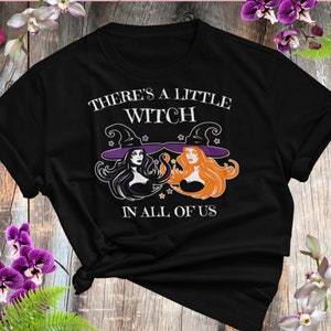 May include: Black t-shirt with a white graphic of two witches wearing purple and orange hats. The text reads "There's a little witch in all of us."