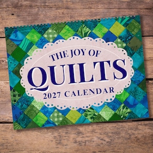May include: A 2027 wall calendar titled The Joy of Quilts. The cover features a diamond-patterned background in various shades of blue and green. Centered is a white scalloped oval label with the text The Joy of Quilts 2027 Calendar in blue.