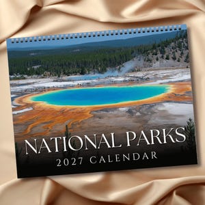 May include: A spiral-bound wall calendar featuring a photograph of the Grand Prismatic Spring with its vibrant blue center and orange-rimmed edges. The text NATIONAL PARKS 2027 CALENDAR is printed in white serif font against a black background at the bottom.