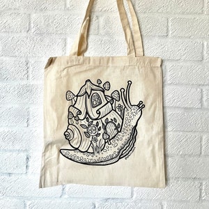 May include: Beige canvas tote bag featuring a whimsical black and white illustration of a snail carrying a house on its back. The snail house has mushroom accents and a door. The tote bag has long beige handles.