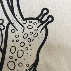 May include: Close-up of a canvas with a black, hand-drawn illustration of a stylized creature. The design features a rounded body with numerous oval-shaped spots and two antennae-like structures.