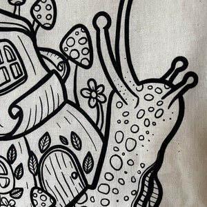 May include: Black and white illustration of a snail carrying a house on its back. The snail's shell is decorated with a whimsical house, mushrooms, and flowers. The design is on a beige fabric.