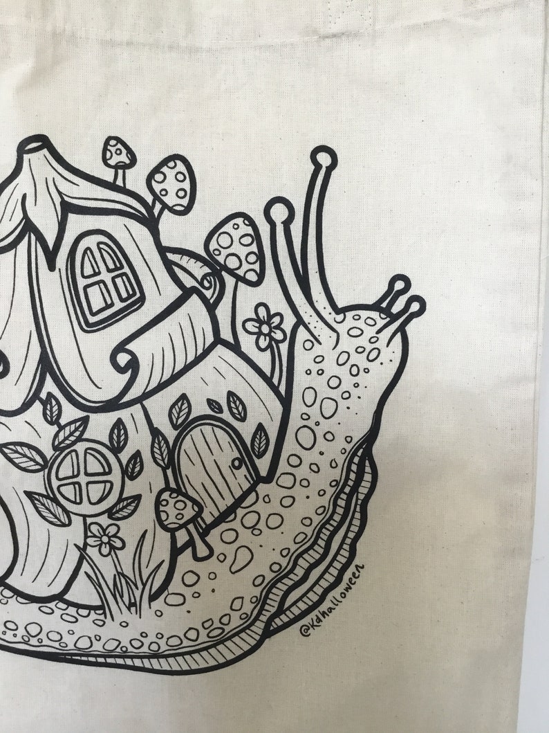 May include: A cream-colored tote bag featuring a whimsical black and white illustration of a snail carrying a house on its back. The snail's shell is detailed with a house, mushrooms, and flowers. The text "@kdhalloween" is in the lower right corner.