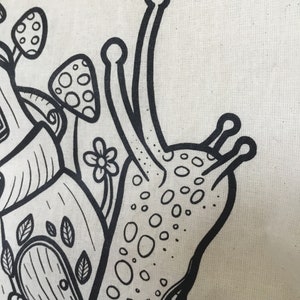 May include: Close-up of a cream-colored tote bag featuring a whimsical black and white illustration. The design includes a snail with a dotted shell, mushrooms, a flower, and a house with a door and leaves.
