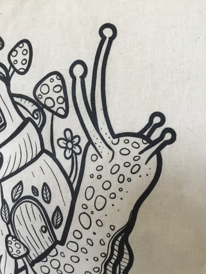 May include: Close-up of a cream-colored tote bag featuring a whimsical black and white illustration. The design includes a snail with a dotted shell, mushrooms, a flower, and a house with a door and leaves.