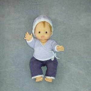 Könnte beinhalten: Eine Strickpuppe, die einen lilafarbenen Strickpullover und eine blaue Strickhose mit weißem Besatz trägt. Die Puppe trägt eine weiße und lilafarbene Strickmütze.