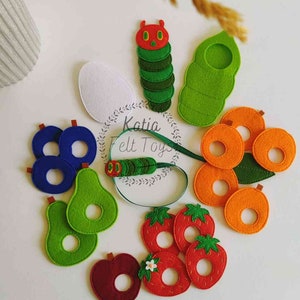 May include: Felt toy set featuring a caterpillar, an egg, a pear, an apple, strawberries, and oranges. The pieces are colorful and have holes in the center. The set is labeled "Katia Felt Toys".