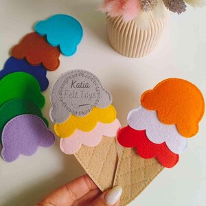 May include: Two felt ice cream cones with different colored scoops. The cones are beige with a quilted pattern. The scoops are red, white, orange, yellow, pink, and gray. The ice cream cones are made by Katia Felt Toys.