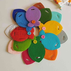 Peut inclure: Un ensemble de poissons en feutre avec des boutons, dans différentes couleurs, dont le rouge, le vert, le bleu, le jaune, l'orange, le violet et le rose. Les poissons sont disposés en cercle, avec le texte "Katia Felt Toys" visible au centre.
