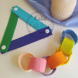 Pode incluir: Uma corrente de feltro colorida com cores do arco-íris, incluindo amarelo, laranja, rosa, roxo e verde. A corrente é feita de anéis individuais de feltro que são costurados juntos. A corrente está sobre uma superfície branca. Katia Felt Toys está escrito em um círculo branco com uma borda verde.