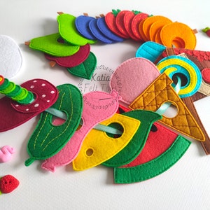 Può includere: Una collezione di forme in feltro colorato che include un uovo bianco, un bruco verde, vari frutti come fragole, pere, arance e anguria, una salsiccia rosa, una fetta di formaggio giallo e un cono gelato, tutti dotati di fori ritagliati.