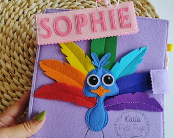 May include: Purple felt book featuring a rainbow peacock design. The book has a pink felt name tag that reads "SOPHIE" and a small circle with the text "Katia Felt Toys".