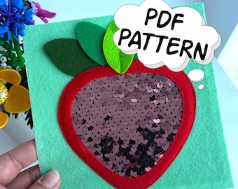 Può includere: Un progetto artigianale in feltro con un quadrato verde con un contorno di mela rosso. Il centro della mela è riempito di paillettes marroni. Foglie verdi in feltro e le parole "PDF PATTERN" in una nuvoletta completano il design.