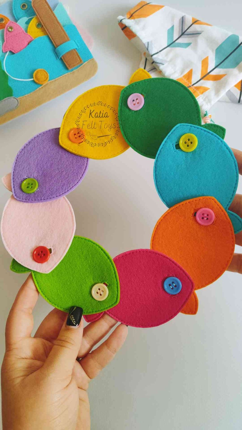 May include: A colorful felt fish garland with different colored fish and buttons. The fish are in a circle and the garland is held by a hand. The text "Katia Felt Toys" is on a yellow fish.