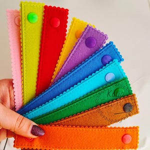 Peut inclure: Un ensemble de bandes de feutre colorées avec des bords festonnés et des boutons. Les bandes sont disposées en arc-en-ciel, avec du rose, du jaune, du rouge, du violet, du bleu, du vert, du marron et de l'orange.