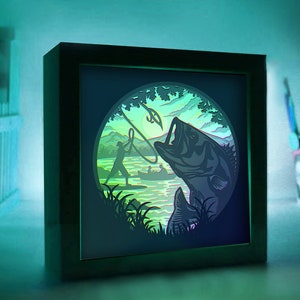 May include: A 3D paper art light sculpture depicting a bass fish jumping out of the water with a fishing hook in its mouth. A silhouette of a person fishing in a boat is visible in the background. The artwork is framed in a black frame.