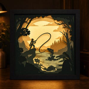 May include: A square, black-framed shadow box featuring a multi-layered paper cutout scene. The artwork depicts a silhouette of a person fishing on a rocky cliff at sunset, with a large sun, pine trees, and a jumping fish in the water below.