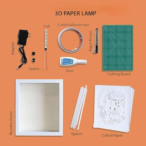 Op de afbeelding: Een 3D-papierlampenset met alle benodigde materialen om een ​​aangepaste lamp te maken. De set bevat een houten frame, een vel gesneden papier, afstandhouders, lijm, een snijmat, een hobbymes, LED-verlichting, een schakelaar, een adapter en dubbelzijdig plakband.