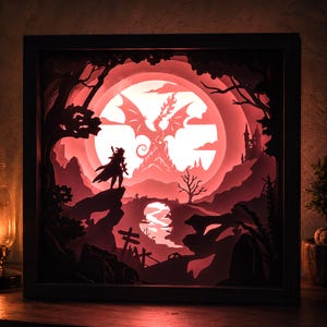 May include: A square shadow box light featuring layered paper cutouts of a fantasy landscape. The scene depicts a silhouette of a warrior standing on a cliff, a large dragon in front of a glowing red moon, a castle, and a winding river below.