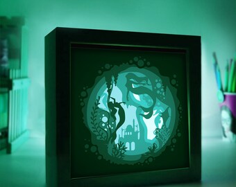 May include: A black framed light-up paper art piece depicting two mermaids swimming in a circle with bubbles and coral. The mermaids are silhouetted against a green background.