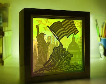 May include: A 3D laser cut wooden shadow box with a patriotic theme. The box features a silhouette of the Statue of Liberty, the US Capitol building, and soldiers raising the American flag. The box is illuminated from behind, creating a glowing effect.