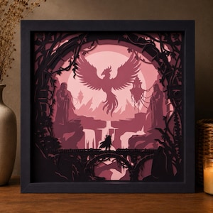 May include: A square black shadow box frame containing a multi-layered paper art piece in shades of pink and dark purple. The scene depicts a central phoenix with wings spread, two standing figures, a bridge, waterfalls, and an arched gateway.