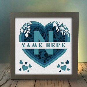 Op de afbeelding: Een wit frame met een blauw en zwart papier gesneden hartontwerp. Het hart heeft de letter "N" in het midden en de tekst "NAME HERE" eronder. Er zijn kleine hartjes verspreid rond het hart.