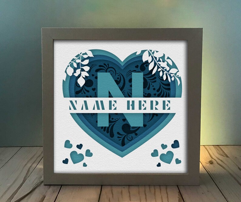 Op de afbeelding: Een wit frame met een blauw en zwart papier gesneden hartontwerp. Het hart heeft de letter "N" in het midden en de tekst "NAME HERE" eronder. Er zijn kleine hartjes verspreid rond het hart.